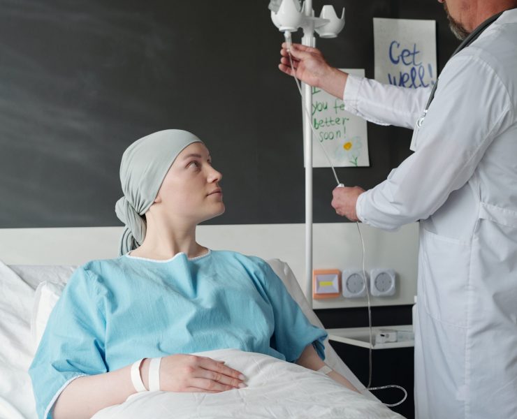 Young female patient with cancer sitting in bed and looking at oncologist preparing dropper for chemotherapy while standing next to woman