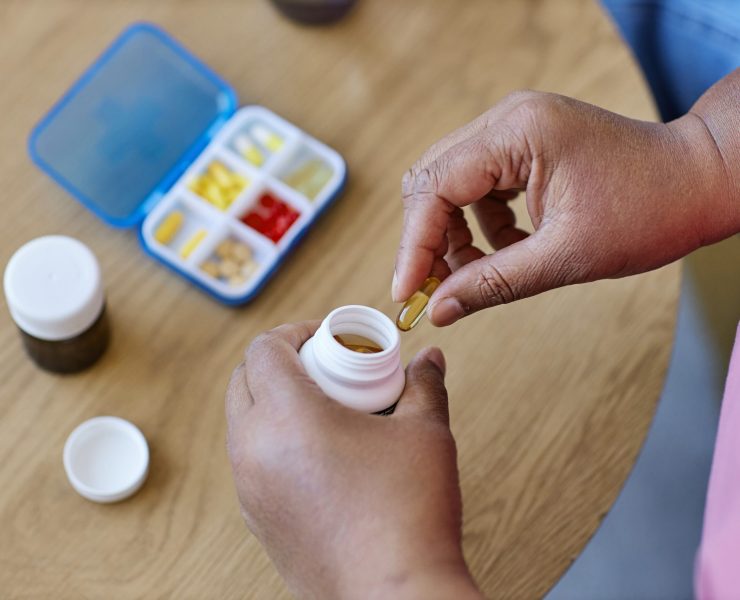 Above view of hands of aged African American woman taking capsule out of white plastic pill-bottle while sitting by table with medicaments