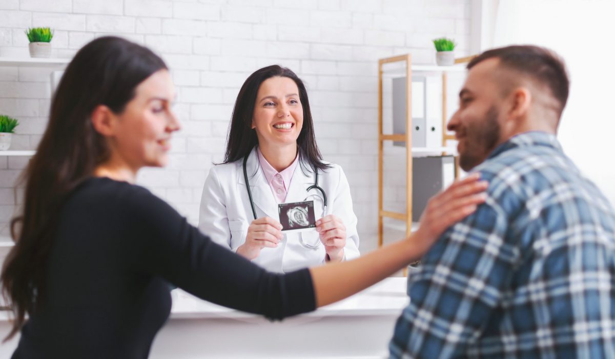 A doctor holds up an ultrasound image of a fetus to a couple sitting across from her in a medical office. The woman is smiling at the image while her partner rests his hand on her shoulder.